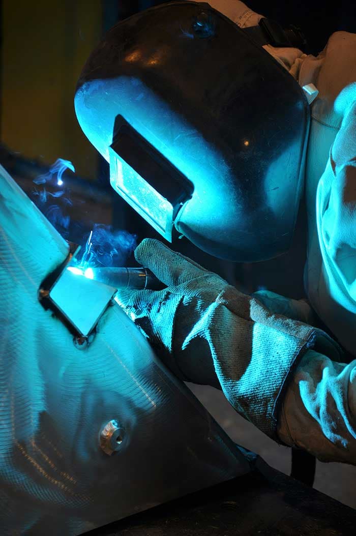 En person sveiser metall mens gnister og blått lys fra sveiseutstyret lyser opp arbeidsområdet